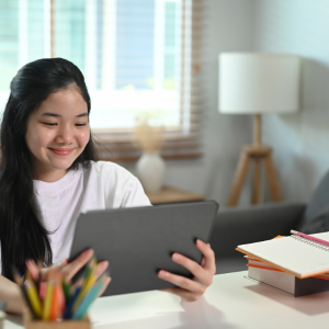 A virtual and hybrid student sits at a desk holding a tablet device. The workspace includes a white lamp, a cup filled with colored pencils, and an open notebook. In the background, there is a window with blinds and some home decor, creating a bright and comfortable study or work environment.